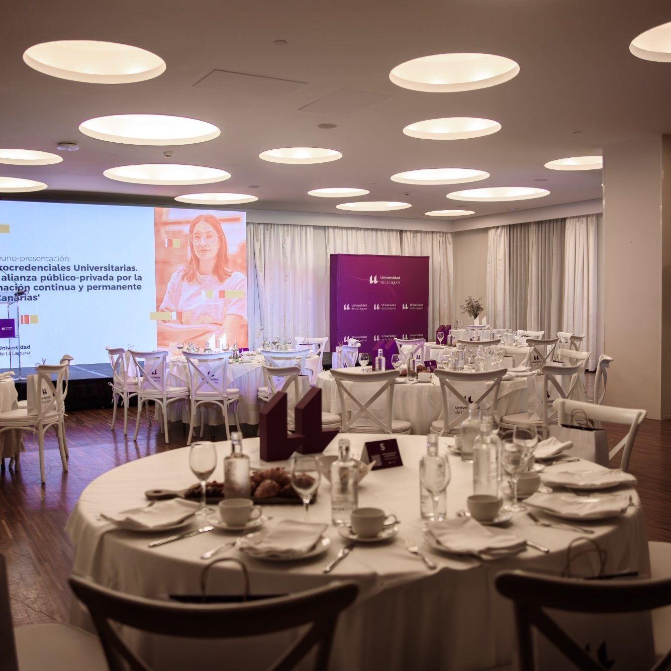 Organización de evento corporativo Sala de eventos con mesas elegantes y proyector en la pared. Decoración blanca y morada.